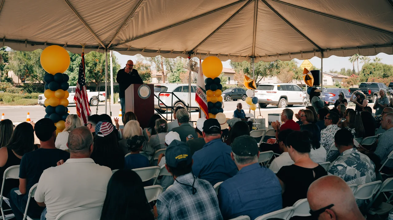 Opening-day guests gathered beneath a canopy, facing a speaker at a podium framed by balloons and flags.
